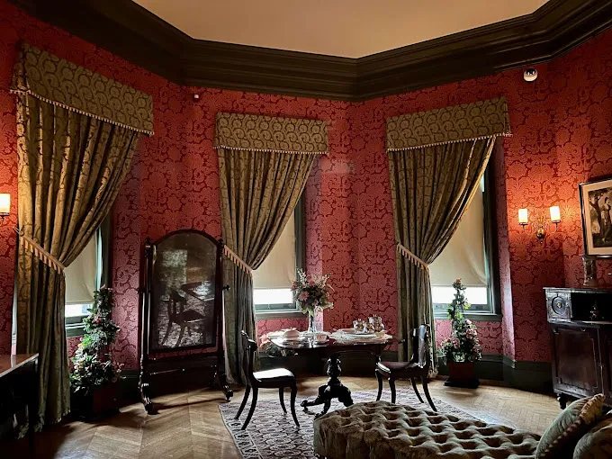 Formal parlor with red patterned walls, draped windows and a polished wood floor