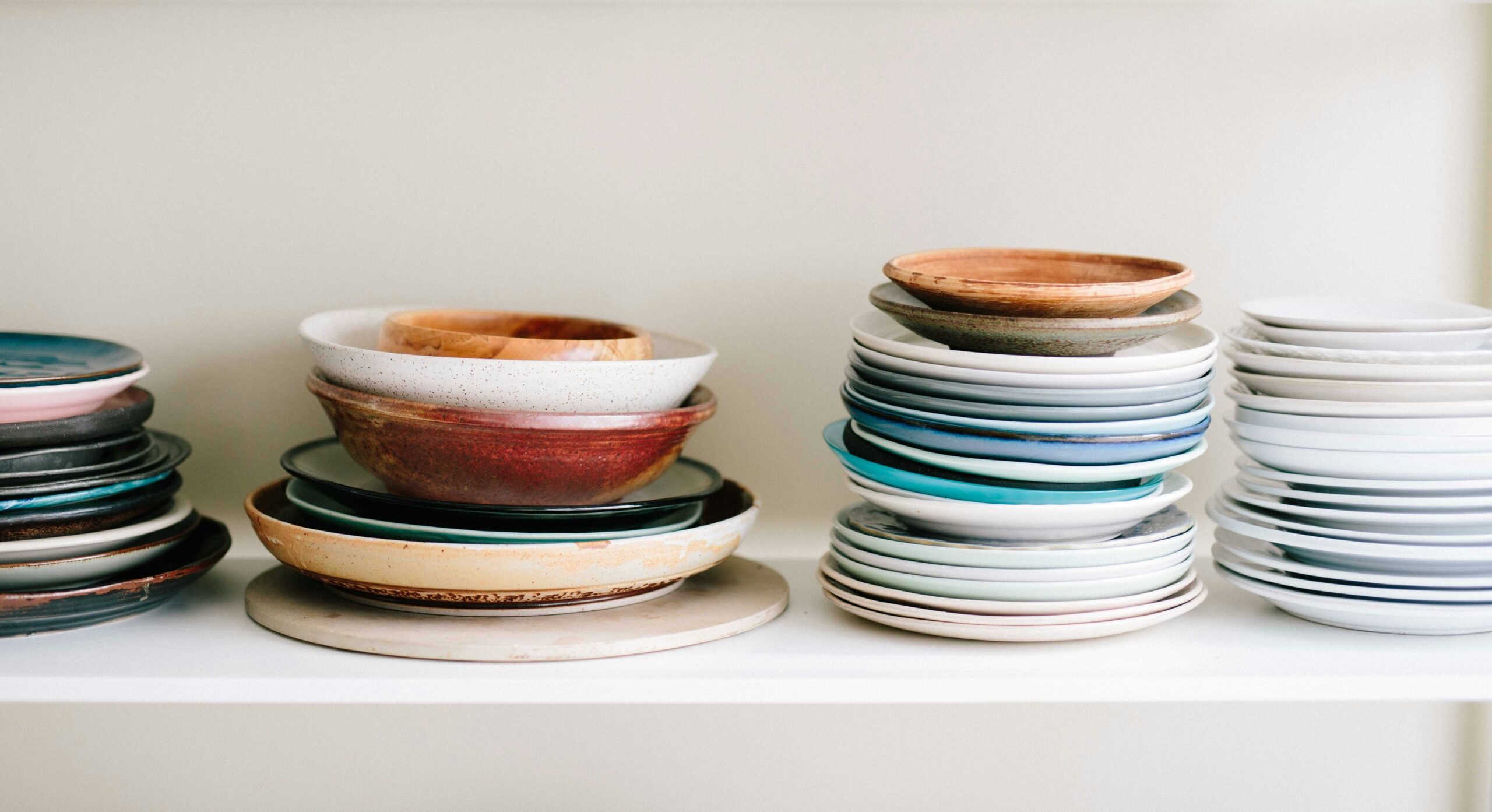 Various stacked ceramic plates and wooden bowls on a white shelf, showcasing a range of earthy and pastel colors, indicative of an organized kitchen cabinet cincinnati ohio