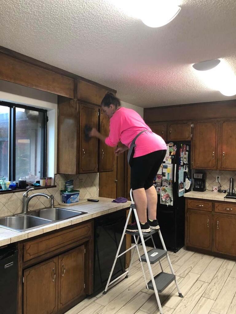 A diligent cleaning specialist wearing a pink shirt and black pants is deep cleaning wooden kitchen cabinets while standing on a step ladder in a Cincinnati home